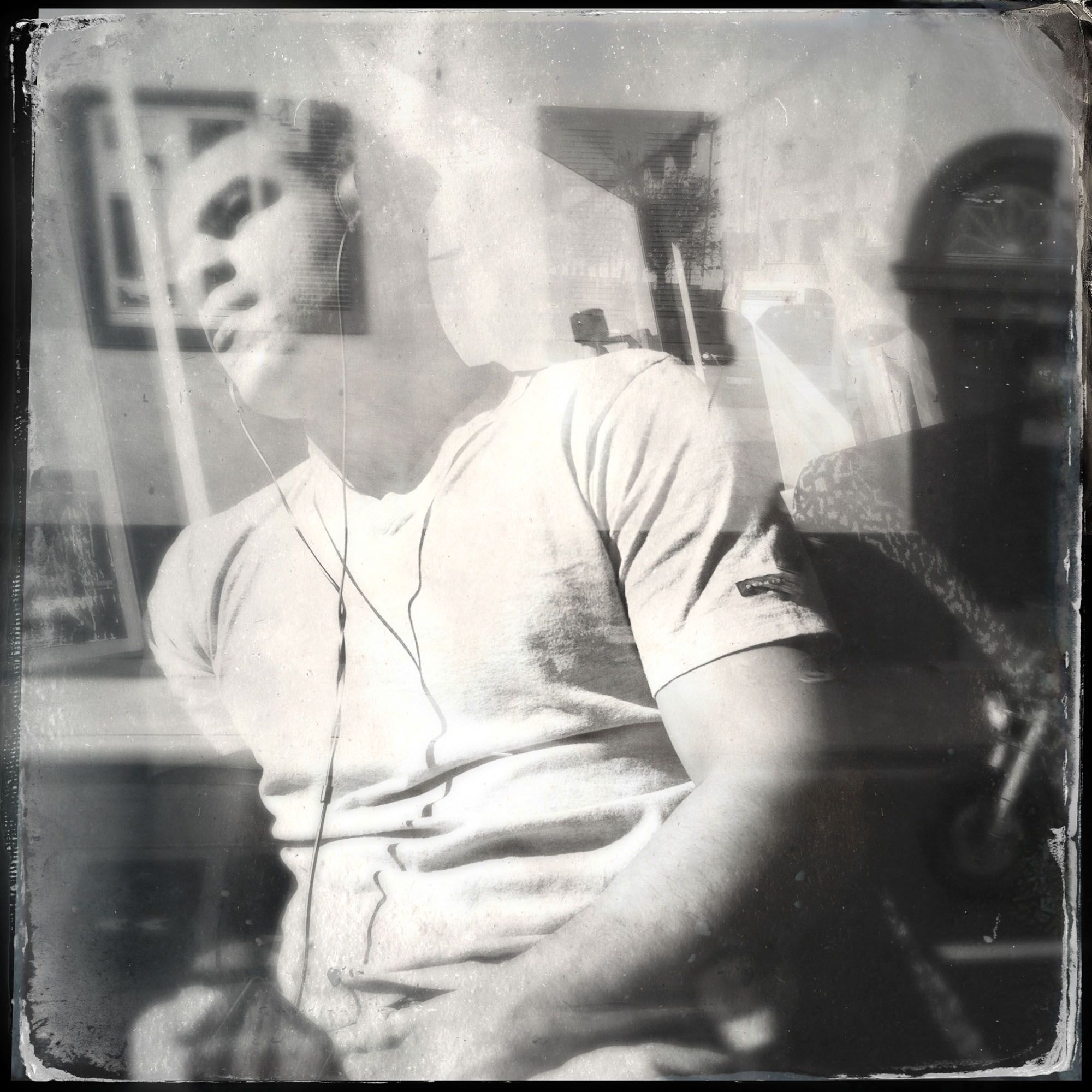 Das quadratische Schwarzweiß-Bild aus der Fotografieserie "Hipstamatic Ghosttrain" des Fotografen Fritz Phillipp aus Frankfurt im Rhein-Main-Gebiet, wurde in Köln aufgenommen und portraitiert einen jungen Mann mit Kopfhörern im Bus, hinter der Doppelbelichtung einer Hausfassade