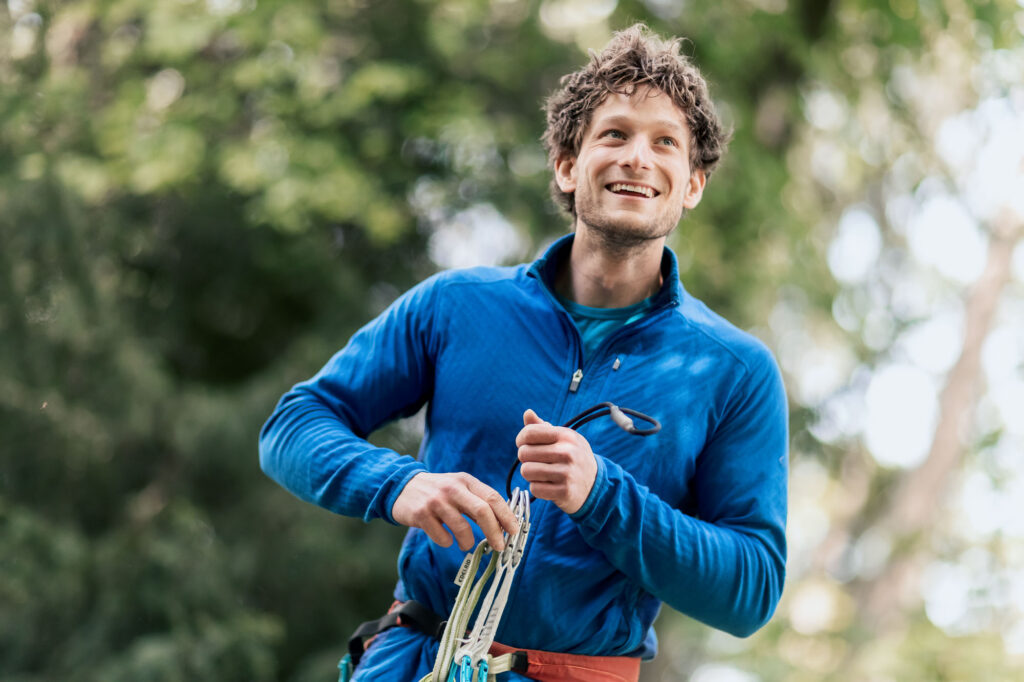 Mann in blauer Outdoor-Kleidung, der lächelnd in die Ferne blickt, während er Kletterausrüstung in den Händen hält. Fotografiert von dem Frankfurter Fotografen Fritz Philipp.