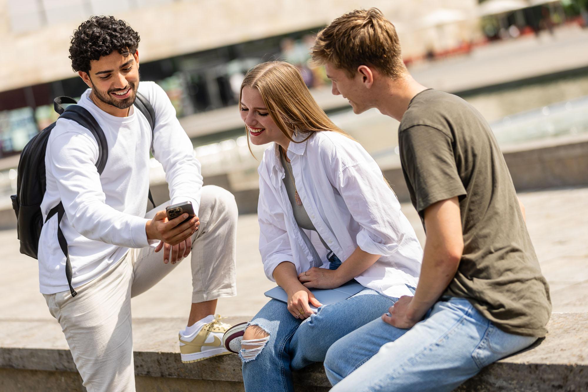 Drei junge Erwachsene sitzen auf einer Steinbank und schauen gemeinsam auf ein Smartphone. Fotografiert von dem Frankfurter Fotografen Fritz Philipp zum Thema visuelle Kommunikation für Bildungseinrichtungen