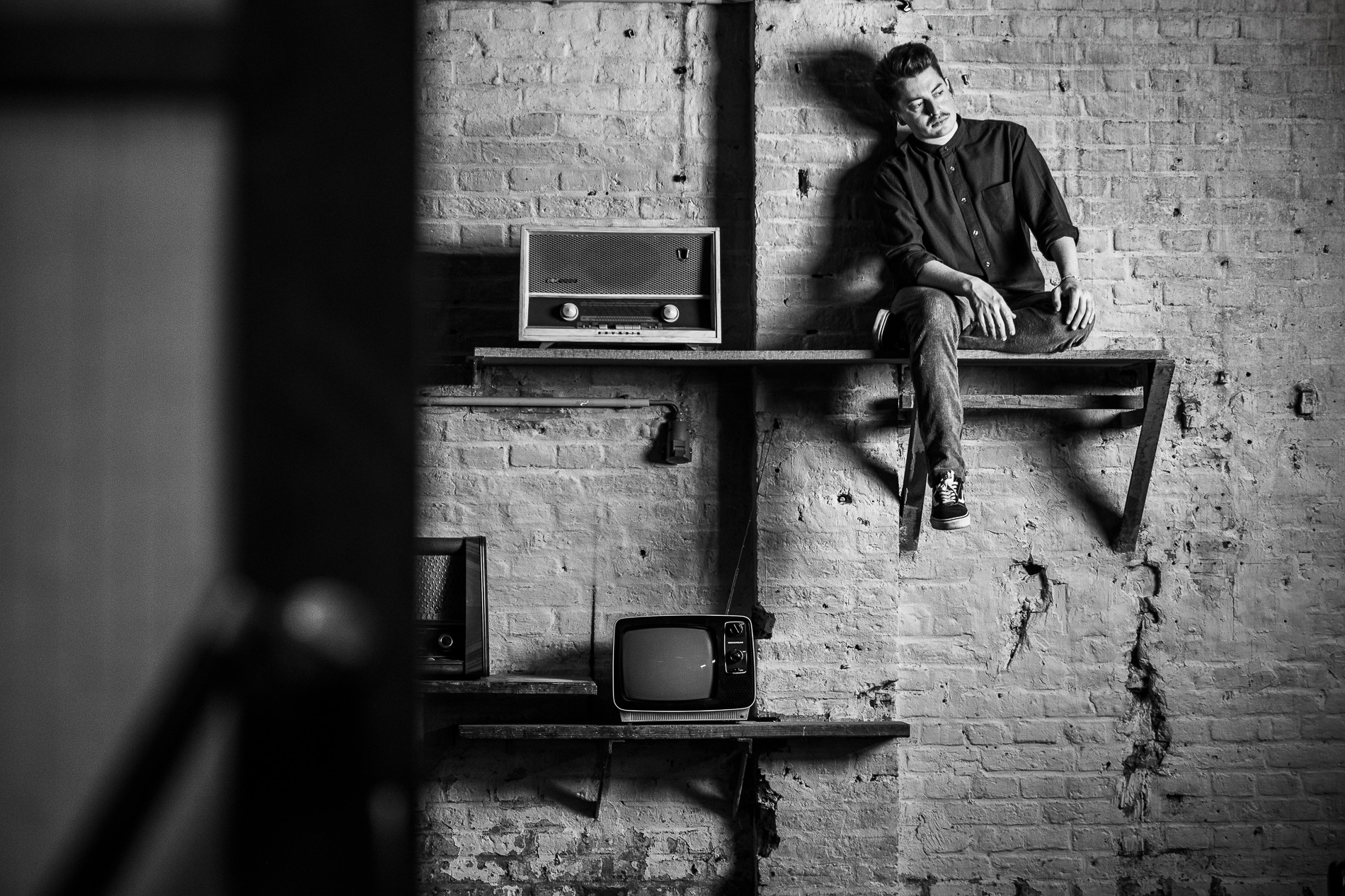 Joscha David Moret sitzt auf einem Wandregal neben einem Retro-Radio und altem Fernseher vor einer Ziegelwand