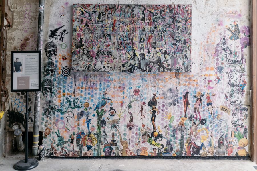 Großformatige Collage mit vielen kleinen Figuren an einer bemalten Wand, davor ein Aufsteller mit Infos zum Künstler – KunstWerkStadt im Schärferaum Bad Homburg