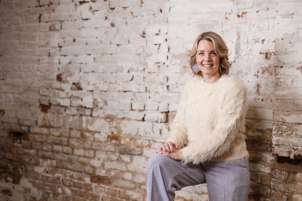 Frau im hellen Strickpullover sitzt lächelnd vor einer weiß getünchten Backsteinwand – Portrait beim Workshop Banking und Generation Alpha im Schärferaum