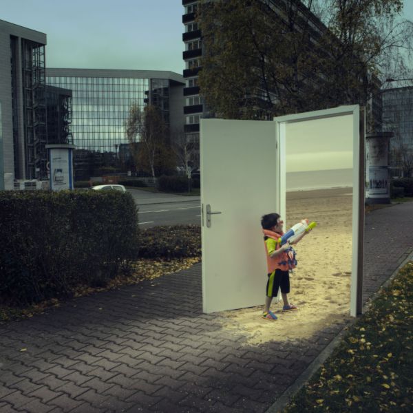 Das surrealistische Foto aus der Serie "Triologie der Türen" für die Darmstädter Tage der Fotografie zeigt einen kleinen Jungen mit Wasserpistole, der durch eine auf einer dunklen Straße stehende Zimmertür auf den sonnigen Sandstrand blickt, von dem Fotografen Fritz Phillipp aus Frankfurt im Rhein-Main-Gebiet