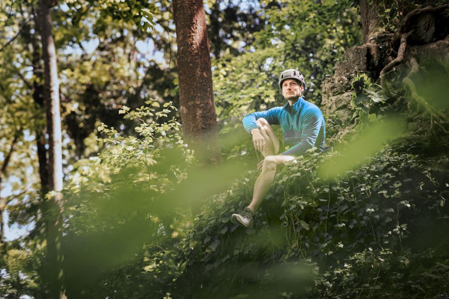 Mann in blauer Outdoor-Kleidung und Helm sitzt auf einem bewachsenen Felsen im Wald, Sonnenlicht fällt durch die Bäume. Fotografiert von dem Frankfurter Fotografen Fritz Philipp.