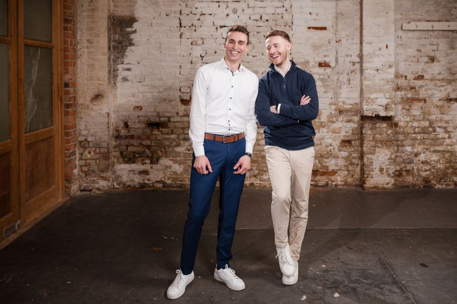 Zwei junge Männer stehen nebeneinander vor Backsteinwand und Holztür im Schärferaum – Portraitshooting Banking und Generation Alpha