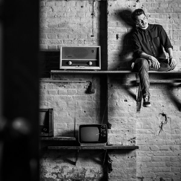 Joscha David Moret sitzt auf einem Wandregal neben einem Retro-Radio und altem Fernseher vor einer Ziegelwand