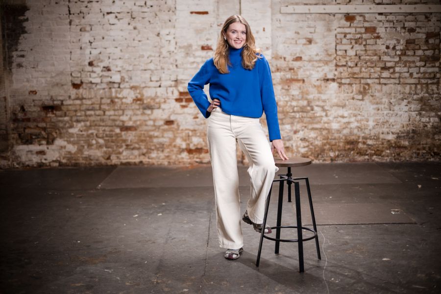 Junge Frau im blauen Pullover lehnt an einem Barhocker vor der Backsteinwand – Portraitshooting Banking und Generation Alpha