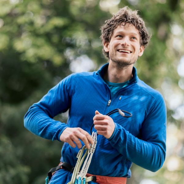 Mann in blauer Outdoor-Kleidung, der lächelnd in die Ferne blickt, während er Kletterausrüstung in den Händen hält. Fotografiert von dem Frankfurter Fotografen Fritz Philipp.