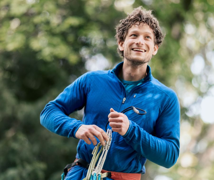 Mann in blauer Outdoor-Kleidung, der lächelnd in die Ferne blickt, während er Kletterausrüstung in den Händen hält. Fotografiert von dem Frankfurter Fotografen Fritz Philipp.
