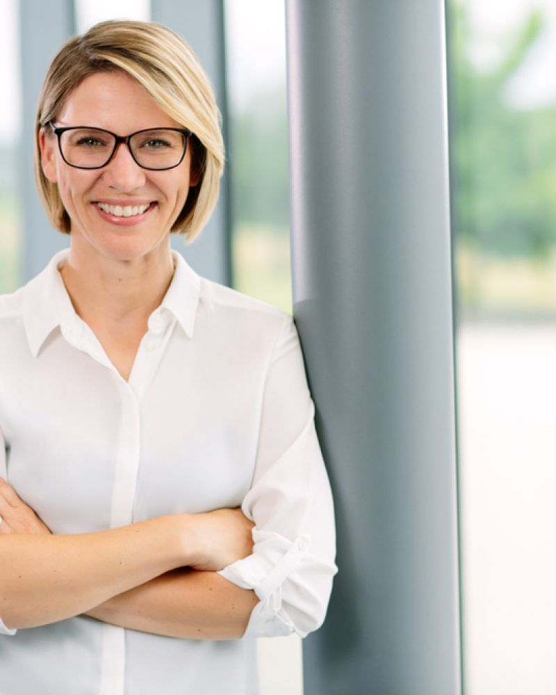 Frau mit Brille und weißer Bluse steht mit verschränkten Armen lächelnd vor einer Glasfront – Businessportrait von Fotograf Fritz Philipp, Bad Homburg