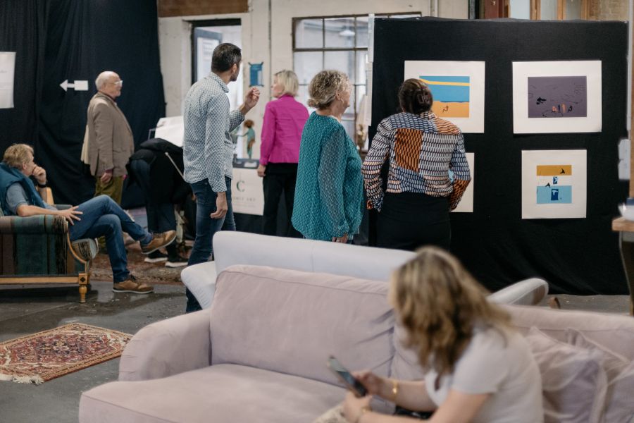 Besucher vor einer schwarzen Stellwand mit gerahmten Grafiken, im Vordergrund ein rosa Sofa – KunstWerkStadt im Schärferaum Bad Homburg